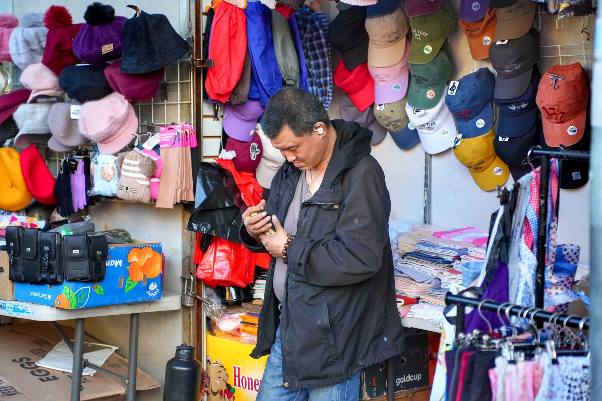 A sidewalk vendor checks his phone beside a dense display of hats and clothing. Nov. 8, 2025.