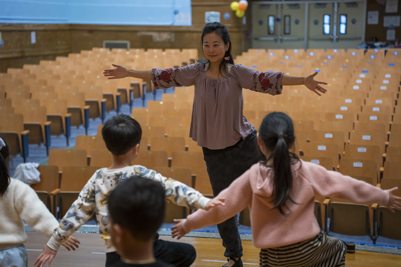 Tang leads from the stage while the children open their arms below.