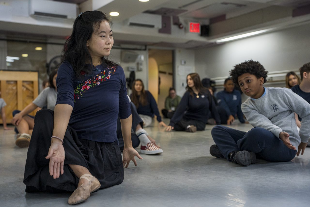 Tang kneels to demonstrate a seated movement while students gather around.