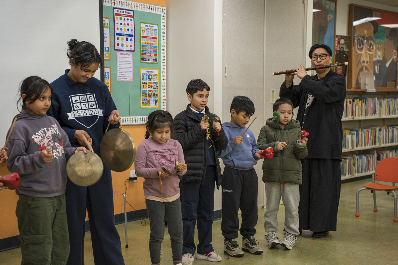 Children join the performance with small percussion instruments as a musician plays flute.