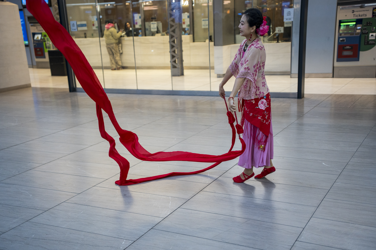 A red ribbon trails across the floor as Tang pauses within the flow of commuters.