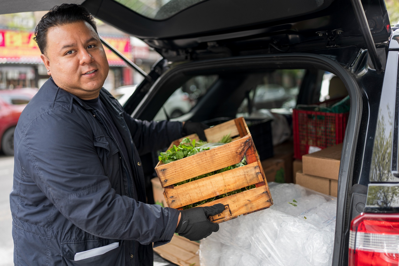 A worker carries a wooden crate of produce beside an open car trunk.